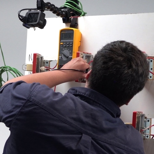 A GoPro mounted to the old troubleshooting board, pointed at the multimeter, while a technician meters the wires behind a device.